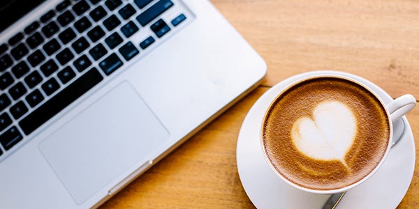 Latte with heart shaped cream sits near laptop before online meeting.